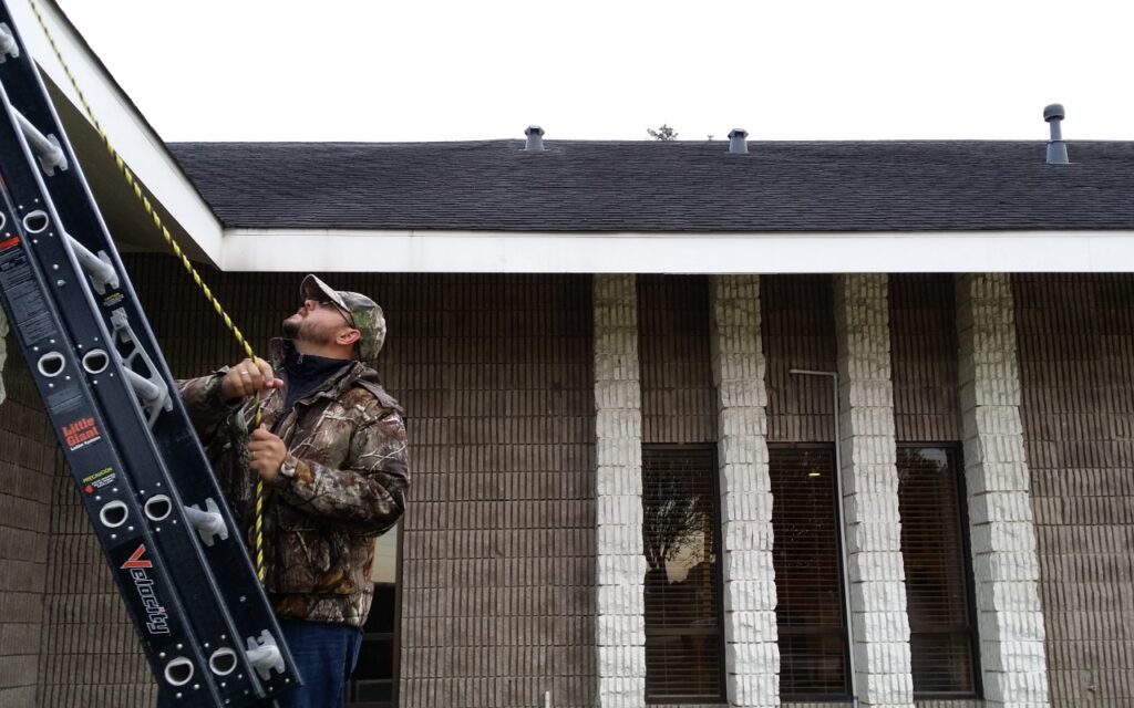 A man in camouflage aims a shotgun outdoors near a building.