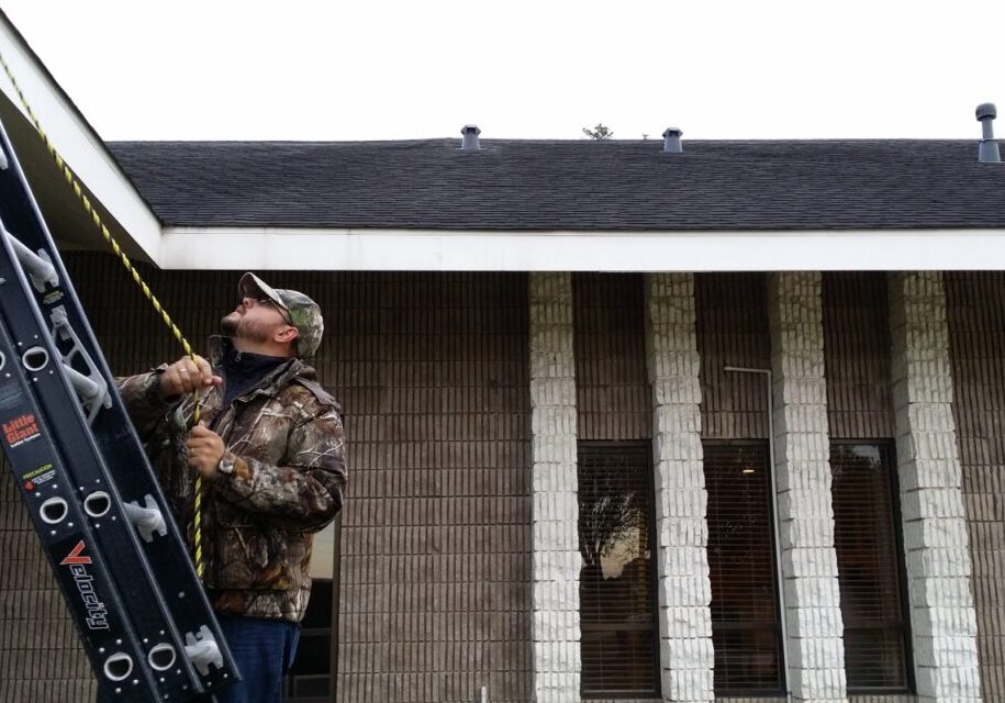 A man in camouflage aims a shotgun outdoors near a building.